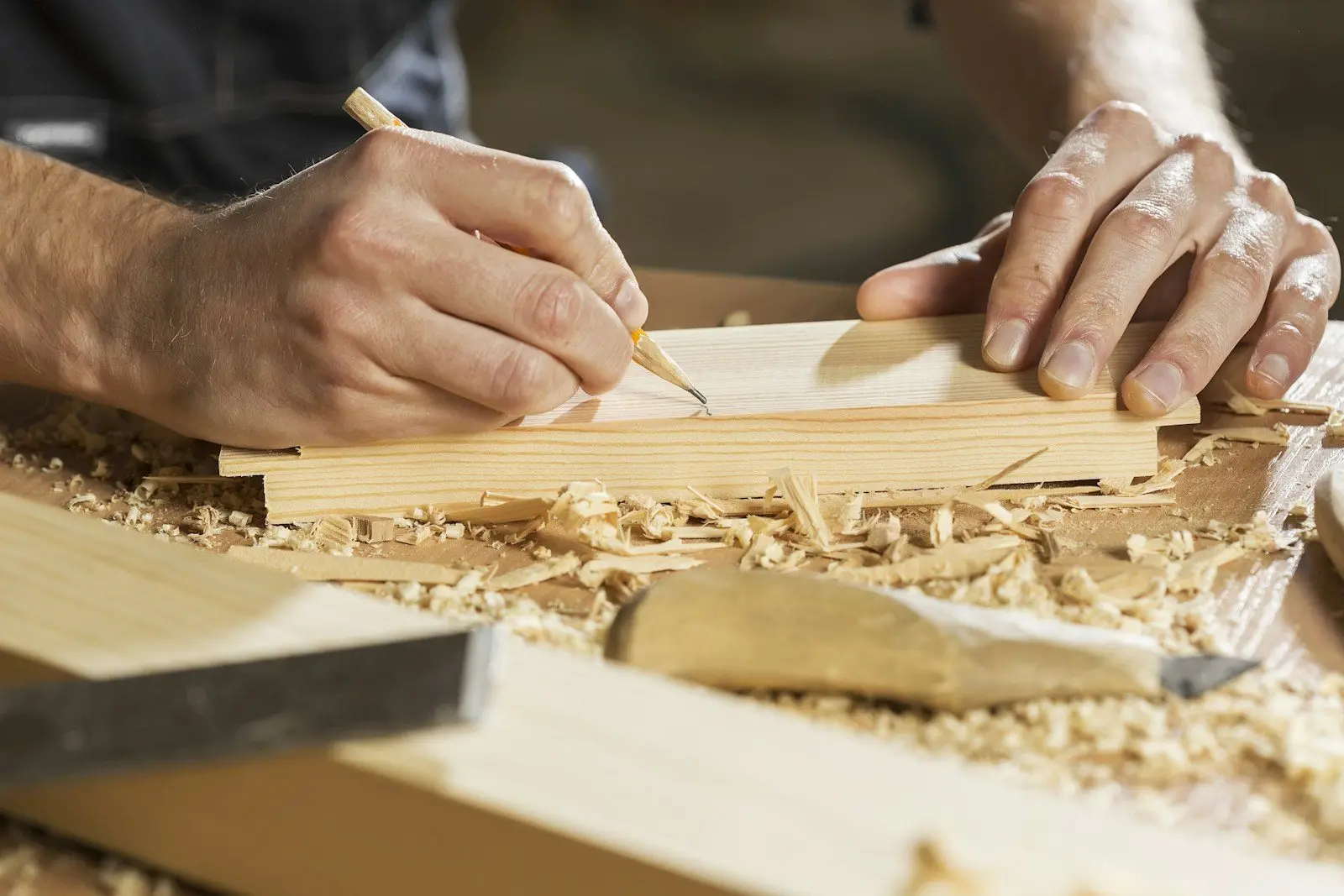 Schreiner reißt mit dem Bleistift ein Maß auf einer Holzleiste an, daneben Hobelspäne